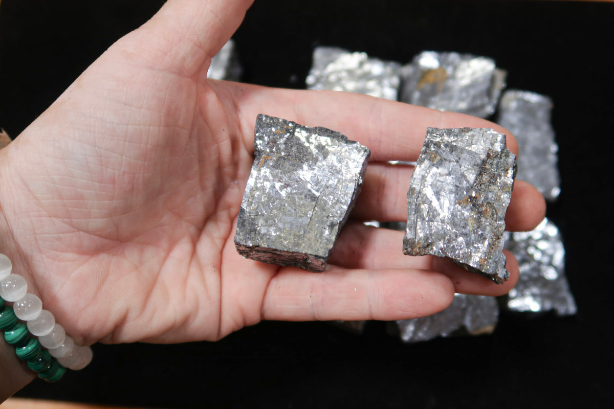 A hand holding two rough, metallic silver mineral or crystal chunks against a black background, with more similar pieces blurred in the background.