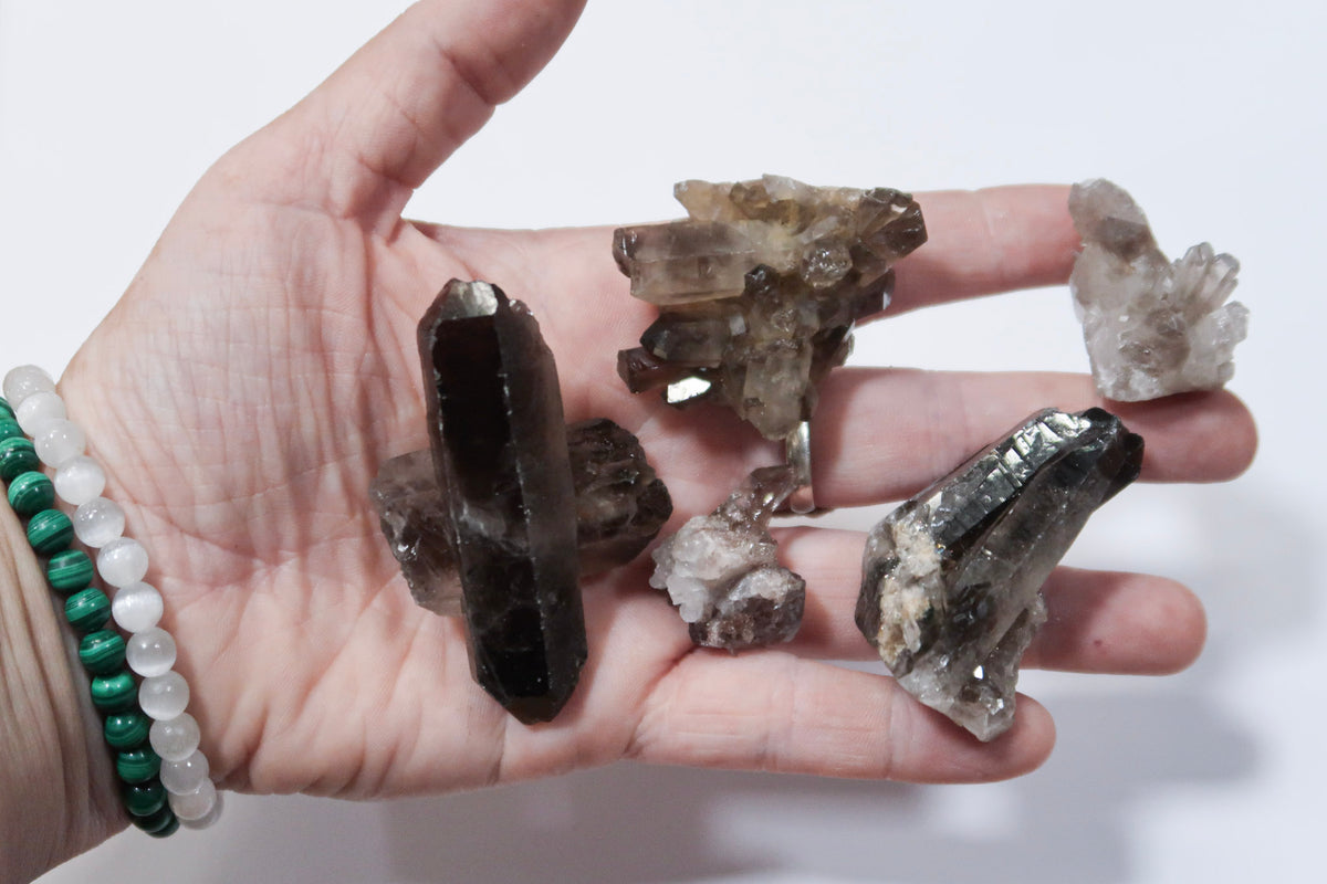 A hand holding several raw smoky quartz crystal clusters and points, with green and white beaded bracelets visible on the wrist, against a plain white background.