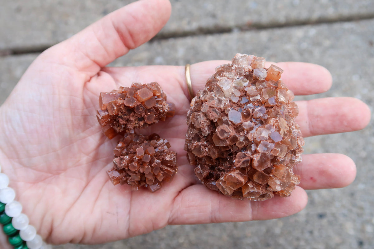 A close-up of a person's hand holding three clusters of brown translucent gemstone crystals against a blurred concrete background.