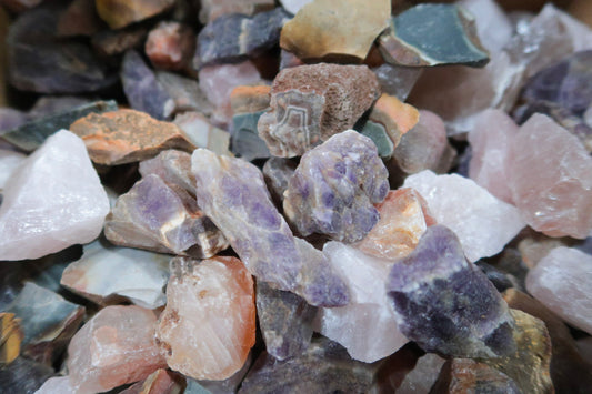 A close-up pile of rough, unpolished crystals and stones in mixed natural colors including purple, pink, white, gray, and brown.